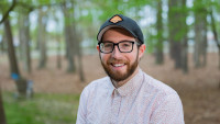 A man wearing a hat poses in front of a natural backdrop. 